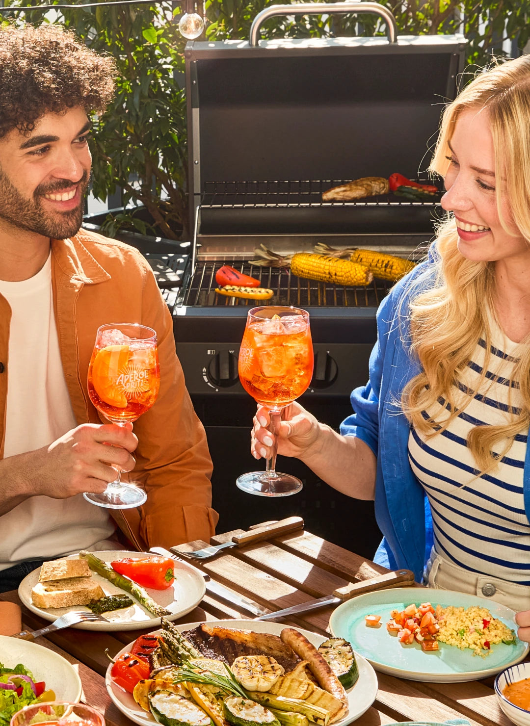 Two friends toasting with Aperol Spritz cocktails at a table filled with grilled food in front of a barbecue